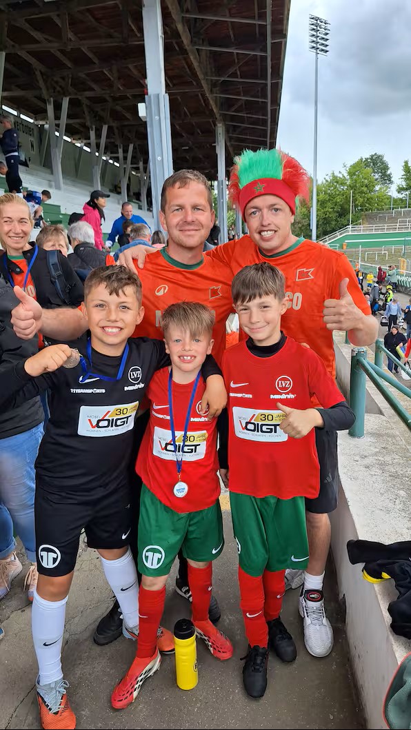 Drei Mini-Kicker des SV Merkwitz mit Medaillen, dazu zwei Trainer im Voigt-Trikot mit Marokko-Fan-Perücke — nach dem Halbfinale der LVZ-Mini-WM 2026 im Alfred-Kunze-Sportpark